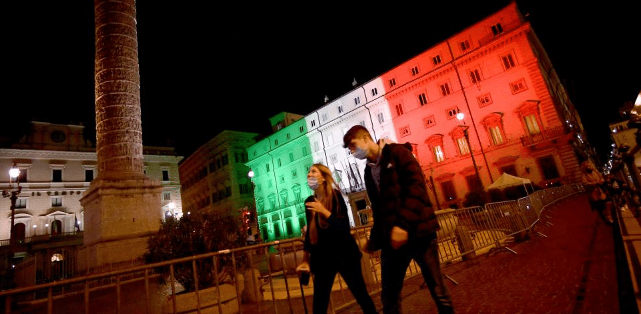 A couple, wearing face masks, walks in front of the Palazzo Chigi Government office in Rome, lit with the color of the Italian flag on November 1, 2020. Credit: AFP Photo