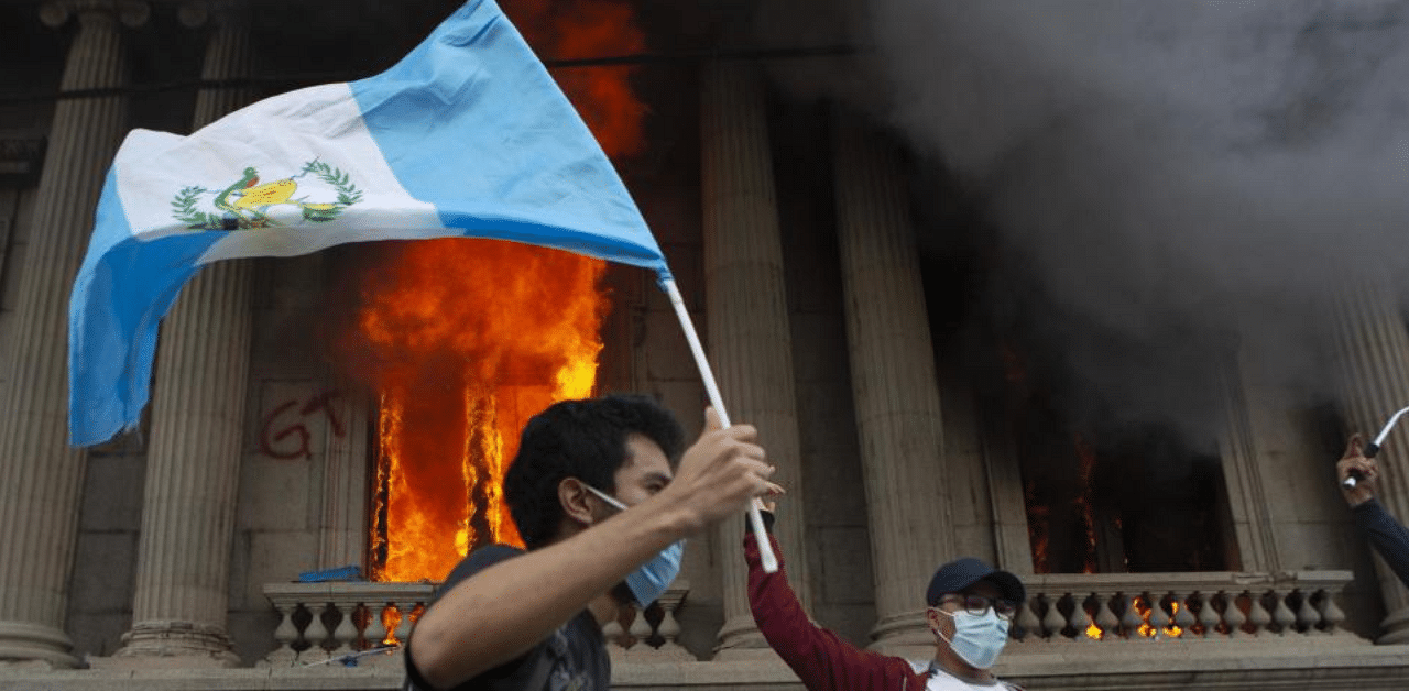 A protester waves a national flag as the Congress building burns in the background, set on fire by protesters, in Guatemala City, Saturday, Nov. 21, 2020. Credit: AP Photo