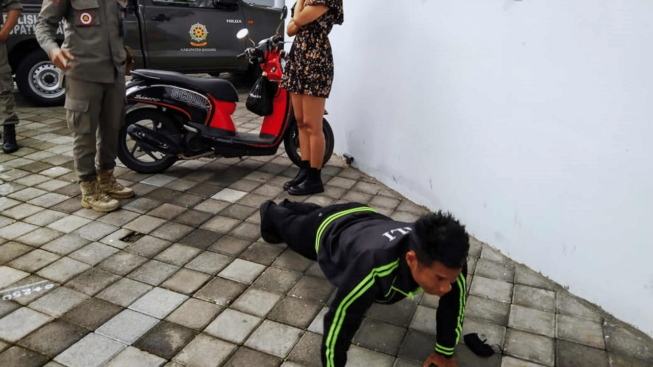 An undated handout picture released on January 20, 2021 by Bali's Satpol PP, the provincial public order agency, shows an official looking on while a man performs push-ups as punishment for not wearing or improperly wearing face masks amid the Covid-19 coronavirus pandemic, along a street in Badung, Indonesia's resort island of Bali. Credit: AFP Photo