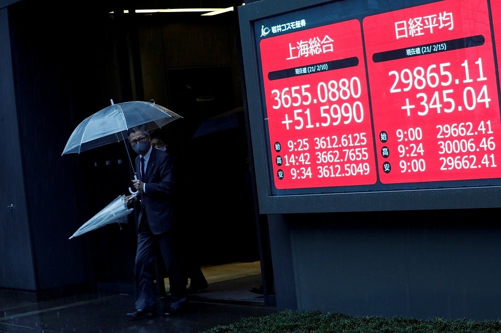 Men holding umbrellas walk near an electric board showing Nikkei index at a brokerage in Tokyo. Credit: Reuters Photo