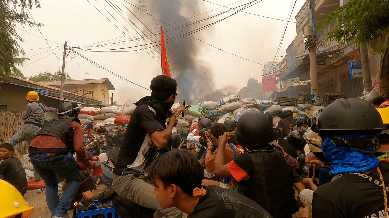 Protesters use slingshots while taking cover behind a barricade as smoke rises from burning debris during ongoing protests against the military coup, in Monywa, Sagaing region, Myanmar. Credit: Reuters Photo