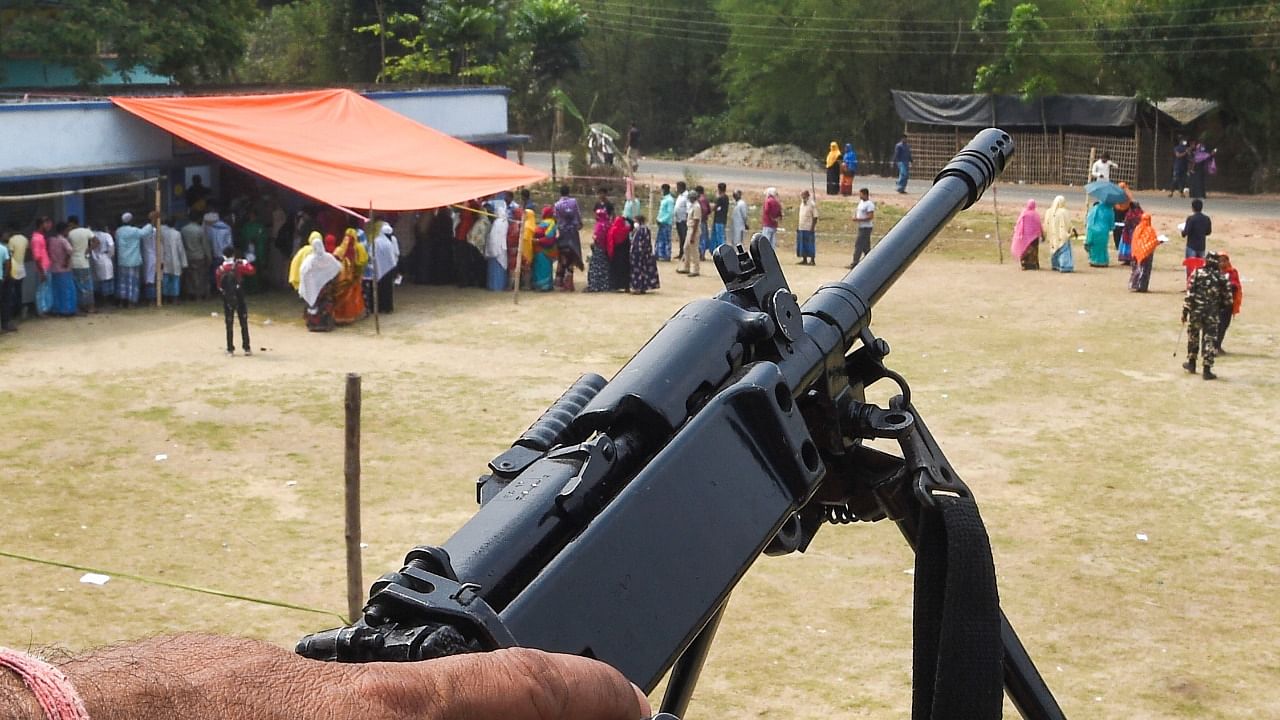 A security person stands guard as people wait to cast their votes at a polling station, during the 4th phase of Assembly elections at Bhangar, in South 24 Pargana, Saturday, April 10, 2021. Credit: PTI Photo