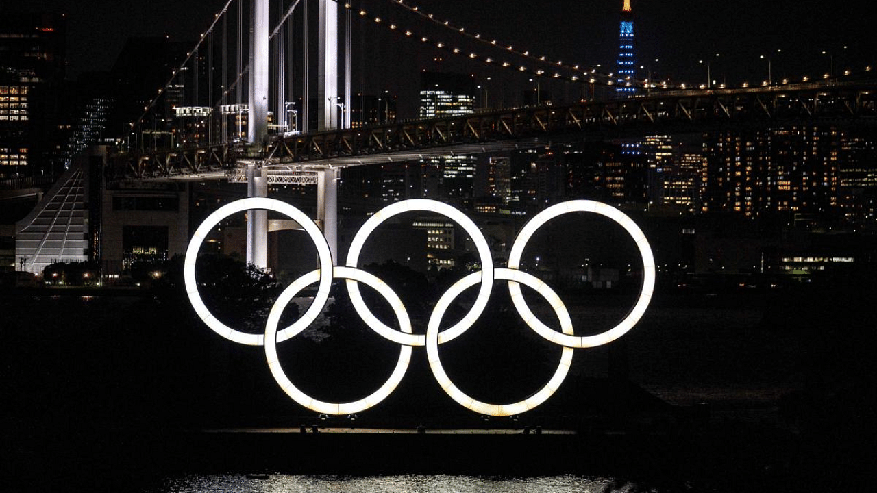 A general view shows the Olympic rings lit up at dusk, with the Rainbow bridge and the Tokyo Tower (back R) in the background, on the Odaiba waterfront in Tokyo. Credit: AFP Photo