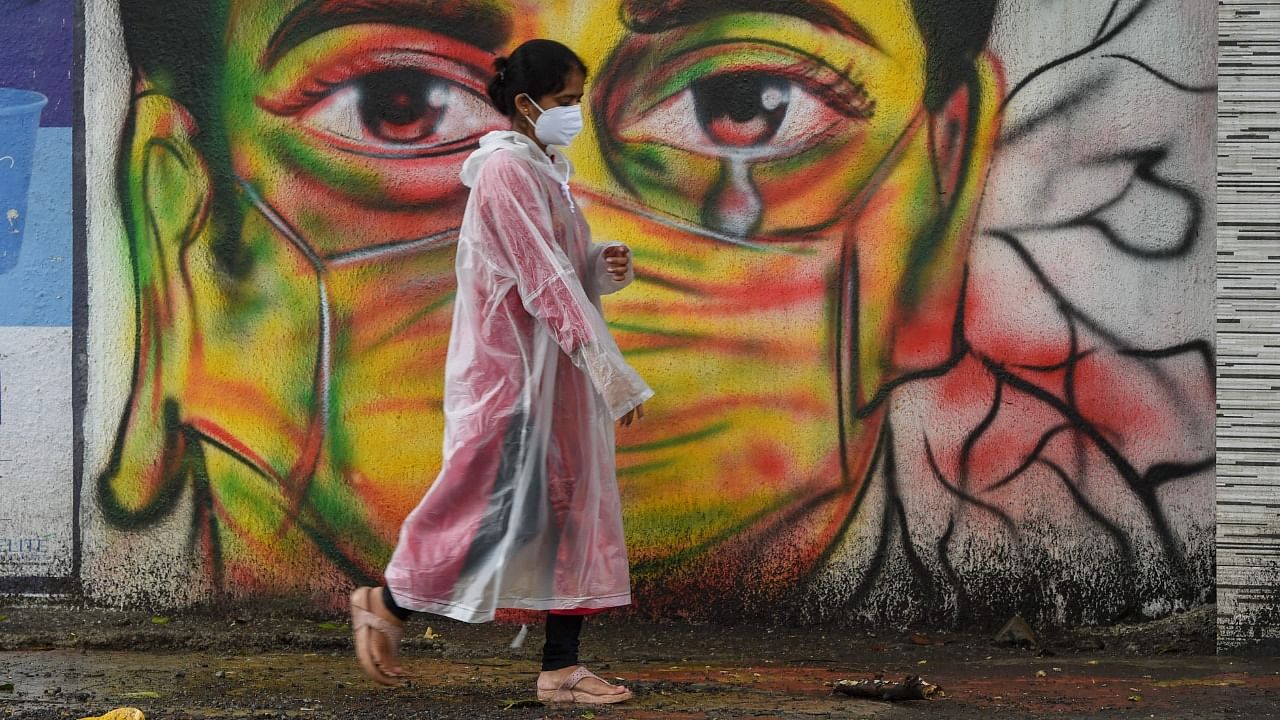 A woman wearing a raincoat walks past a mural depicting a woman with a facemask to spread awareness about the Covid-19 coronavirus, in Mumbai on June 17, 2021. Credit: AFP Photo