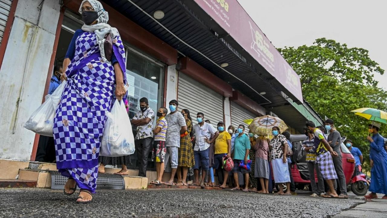 A woman carrying food bags walks pasts people standing in queue outside a state-run supermarket to buy essential food items in Colombo. Credit: AFP Photo