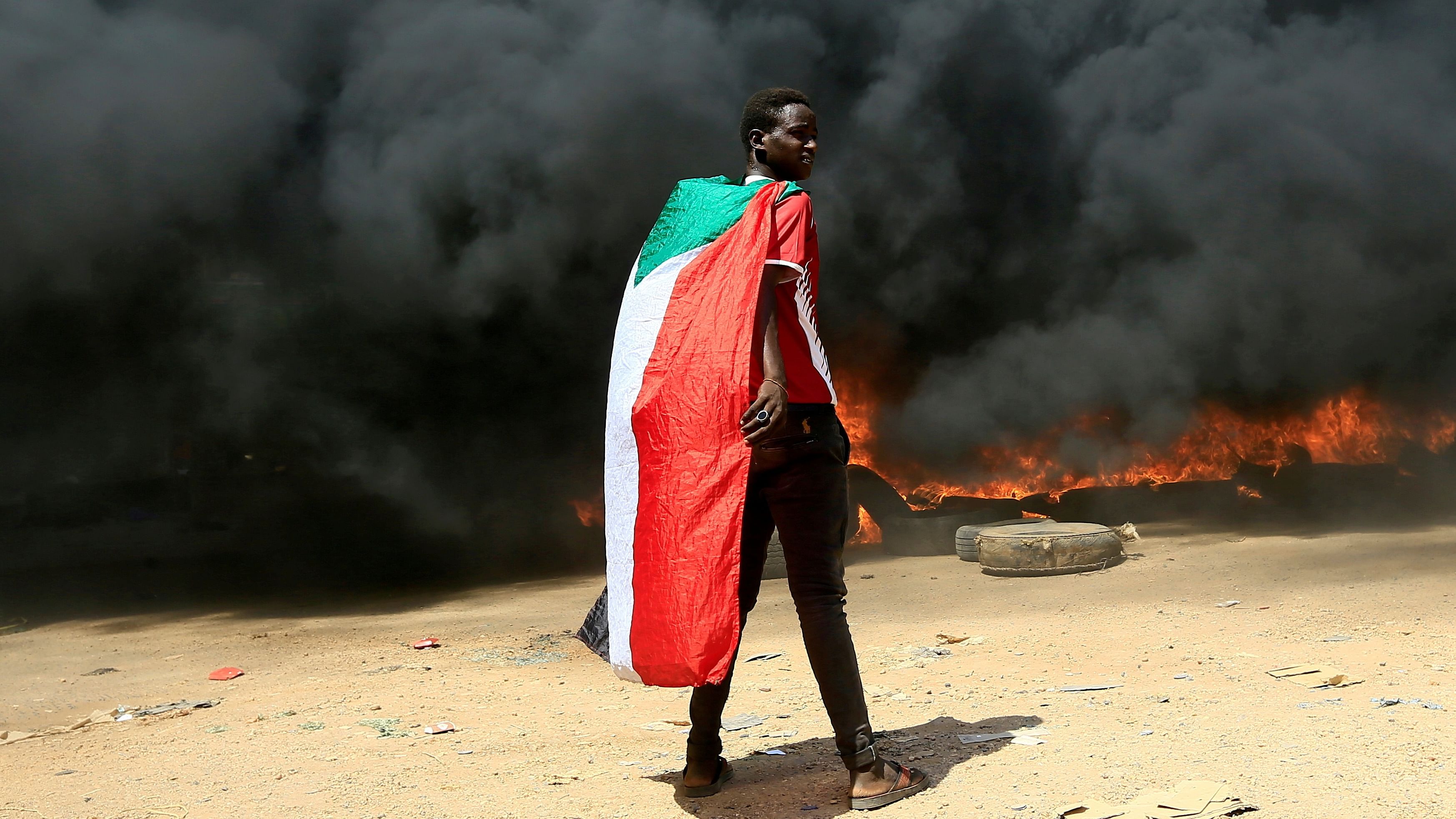 A person wearing a Sudan's flag stand in front of a burning pile of tyres during a protest against prospect of military rule in Khartoum, Sudan. Representative image. Credit: Reuters File Photo