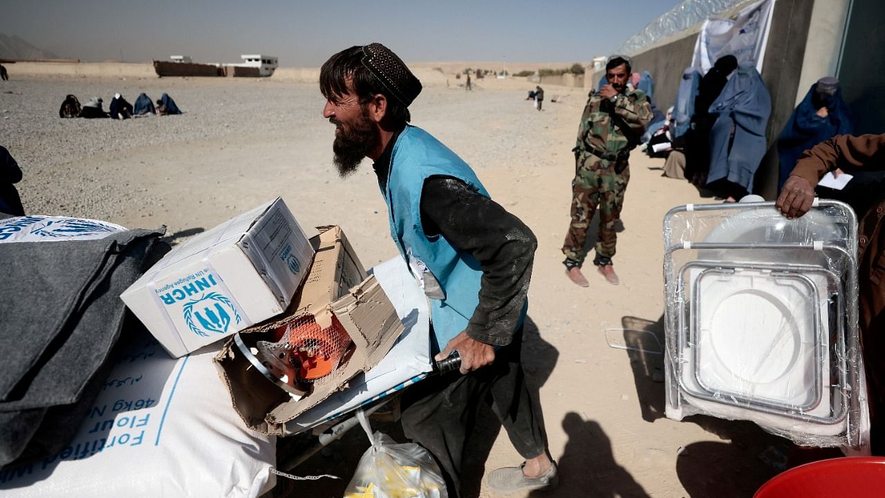 A UNHCR worker pushes a wheelbarrow loaded with aid supplies for a displaced Afghan family outside a distribution center as a Taliban fighter secures the area on the outskirts of Kabul, Afghanistan. Credit: Reuters File Photo