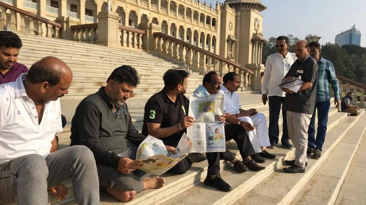 KPCC President DK Shivakumar and Congress MLAs sitting on the steps at Vidhana Soudha premises and reading daily newspaper during their protest. Credit: IANS Photo