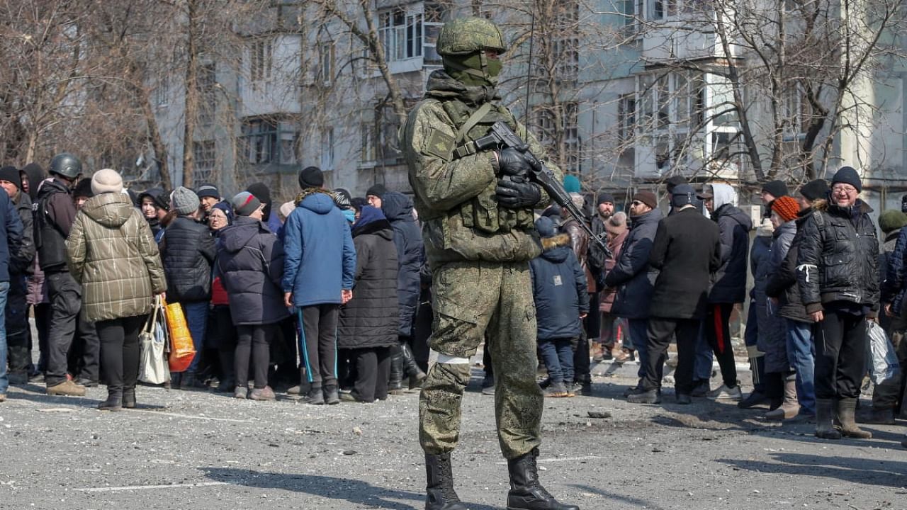 A Russian army soldier stands next to local residents who queue for humanitarian aid delivered during Ukraine-Russia conflict, in the besieged southern port of Mariupol. Credit: Reuters photo