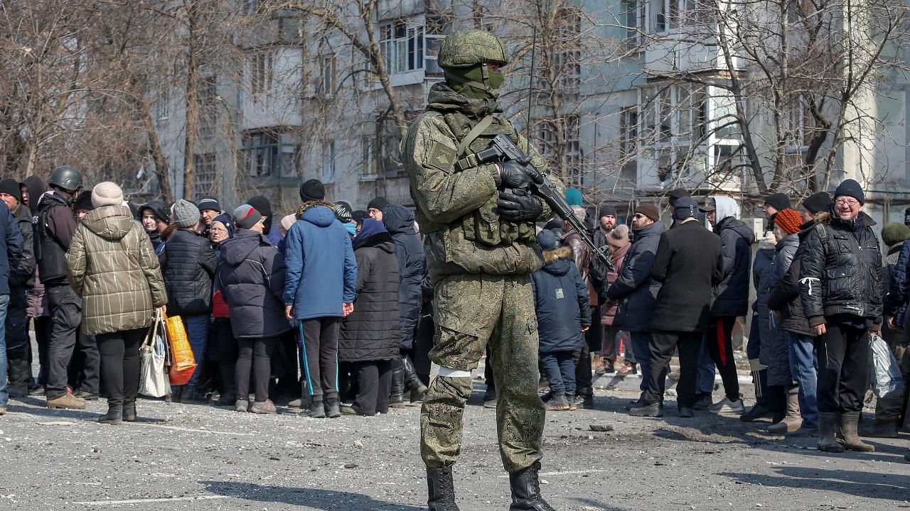A Russian army soldier stands next to local residents who queue for humanitarian aid delivered during Ukraine-Russia conflict, in Mariupol. Credit: Reuters File Photo
