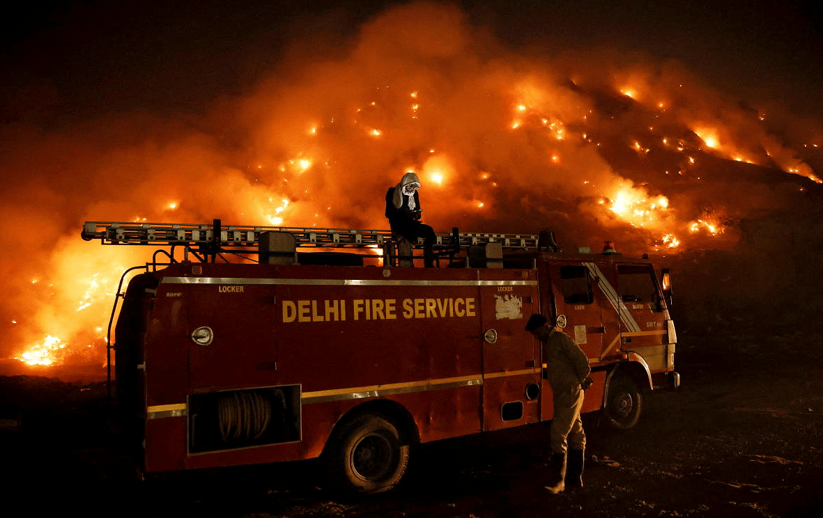 A  firefighter uses his mobile phone as he sits on top of a fire truck as smoke billows from burning garbage at the Bhalswa landfill site in New Delhi. Credit: Reuters