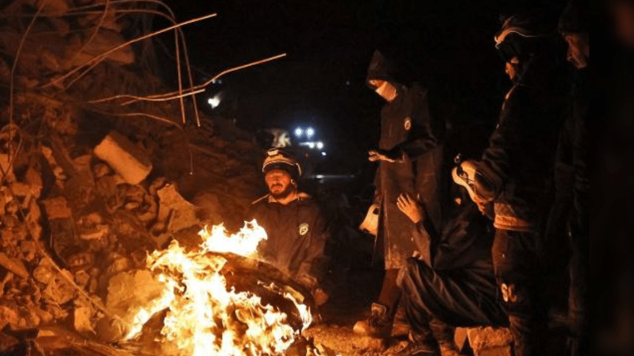 White Helmet Syrian rescue workers warm themselves by a fire next to a collapsed building late on February 6, 2022 in the town of Sarmada, in Syria's rebel-held northwestern Idlib province. Credit: AFP Photo.