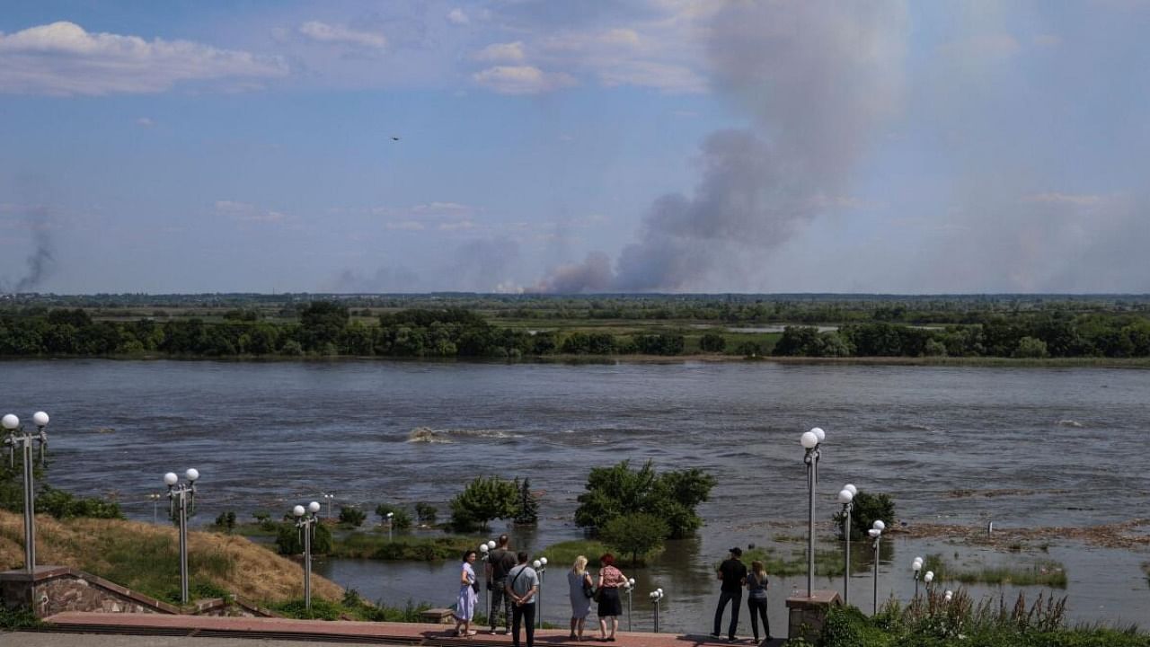 Locals near the Dnipro river which flooded after the Nova Kakhovka dam breached as smoke rises after shelling on the opposite bank. Credit: Reuters Photo