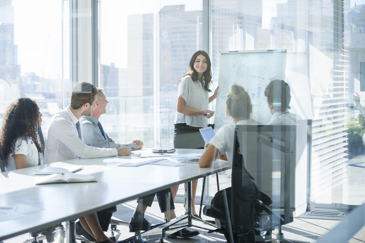 Woman giving a presentation to her team. She is using a whiteboard with charts and graphs. She is talking. They are in an office boardroom at the table with laptop computers and paperwork. She is smiling. Men and women in the group.art