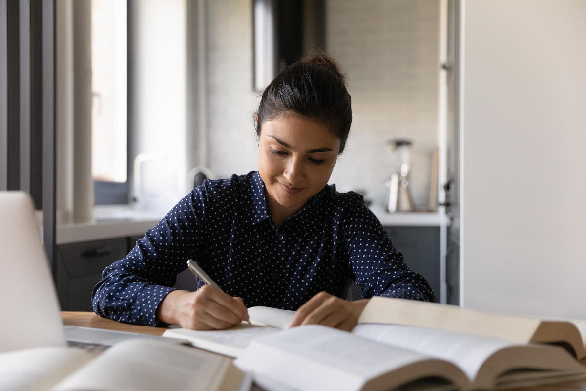 Close up Indian woman writing notes or essay, studying, reading textbook, smiling student sitting at table with books, preparing for university or college exams, doing homework, school assignmentsClose up Indian woman writing notes, studying, reading tex