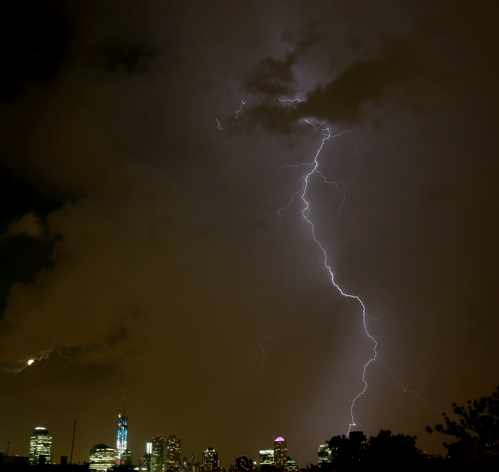 With One World Trade Center, third from left, dressed in red, white and blue lights and a moon in its waning gibbous stage peeking through clouds, left, a lightning bolt from a thunderstorm strikes down over Lower Manhattan Tuesday, June 25, 2013, as...