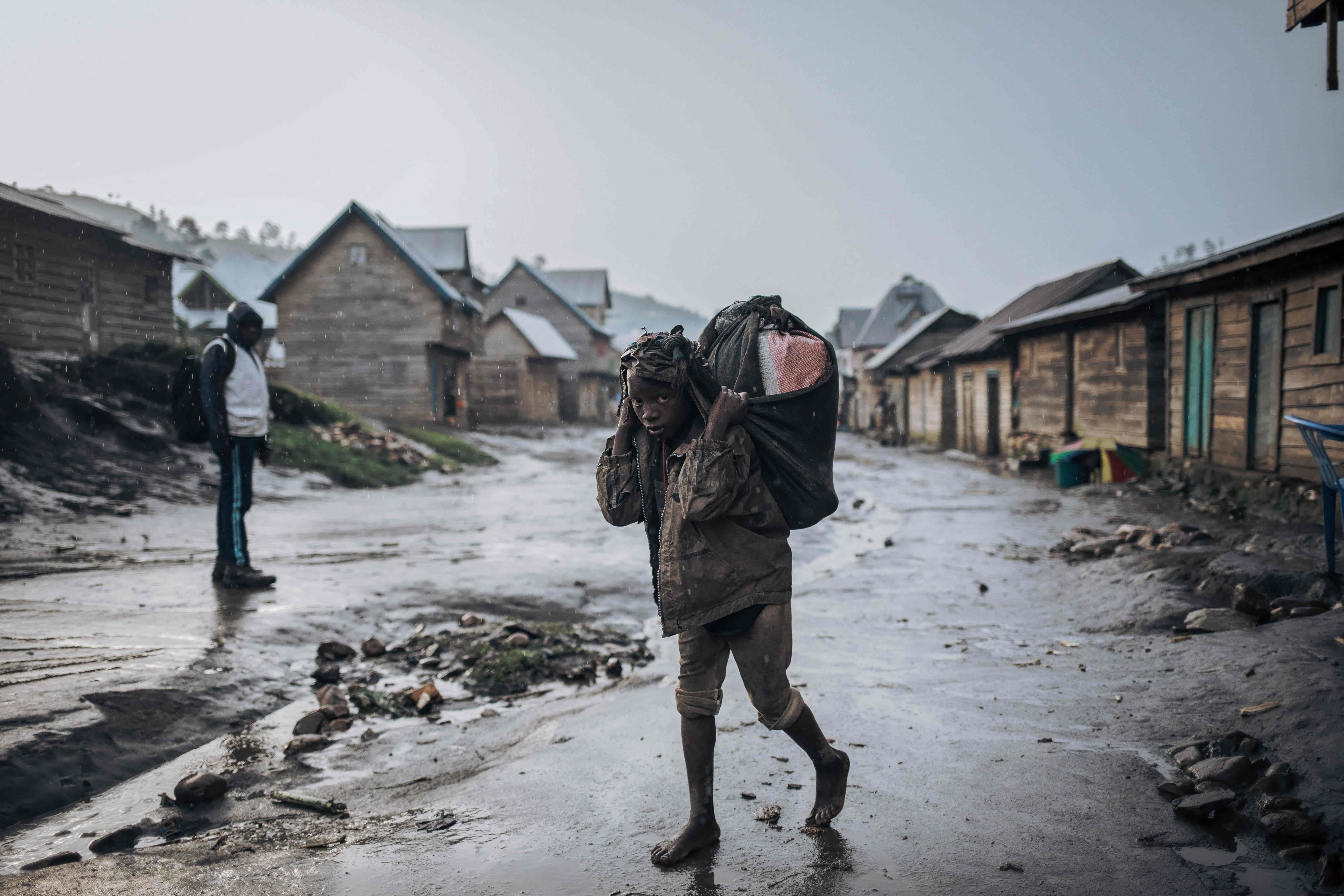 A child carries goods on his back in the rain on the main street of Muheto, a three-hour motorcycle ride from Masisi Centre in the mountains where armed groups regularly attack, on March 28, 2022, in North Kivu province, eastern Democratic Republic of Congo. Credit: AFP Photo